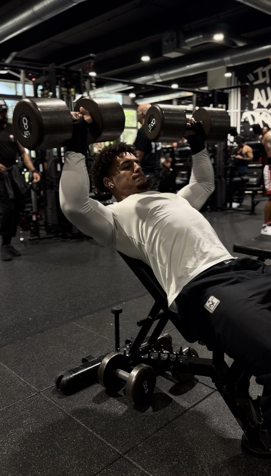 Eli Hughes performing an incline dumbbell press with heavy weights inside a Miami gym