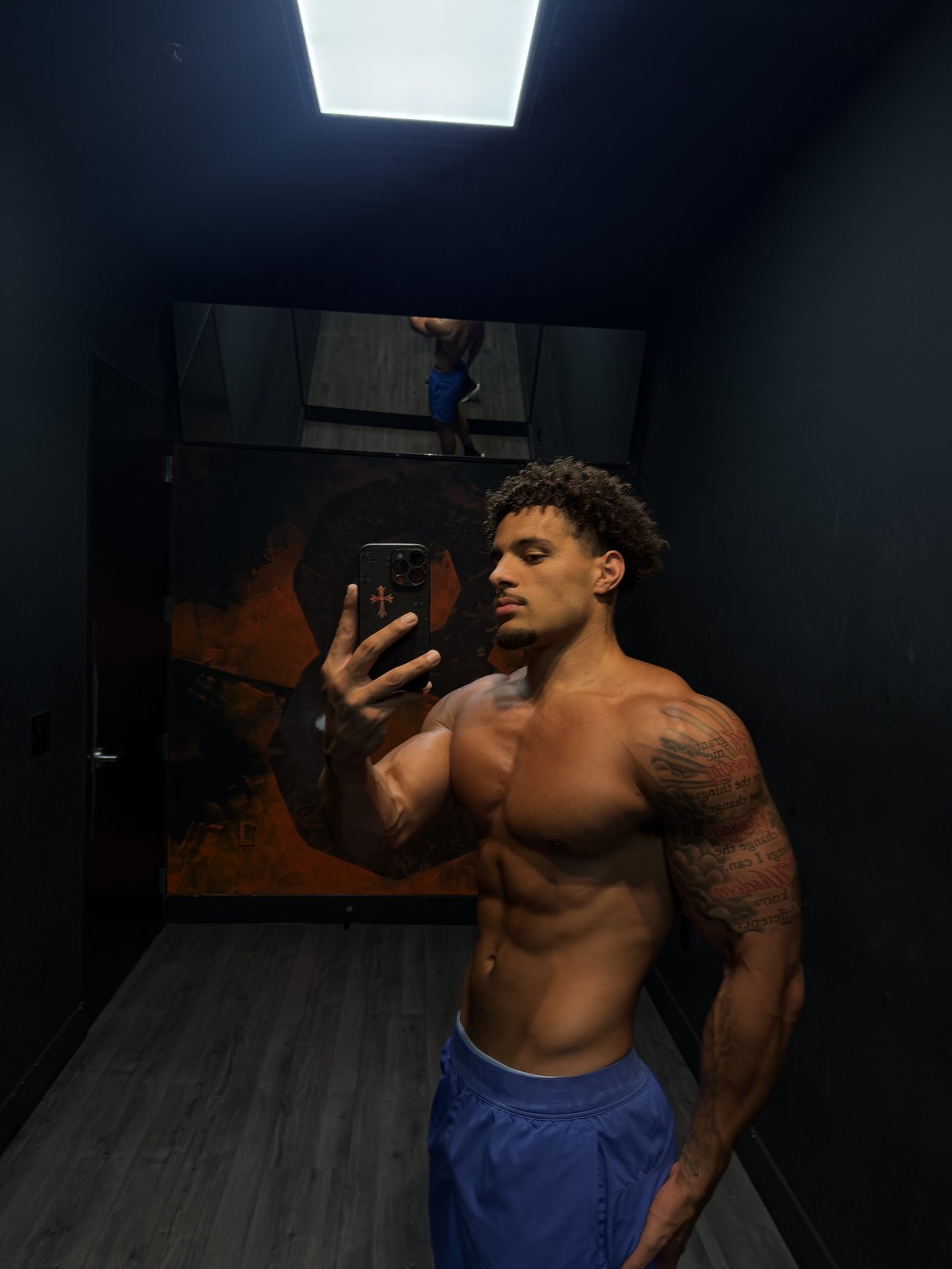 Eli Hughes flexing in a dimly lit gym mirror selfie, showing his tattoos and physique