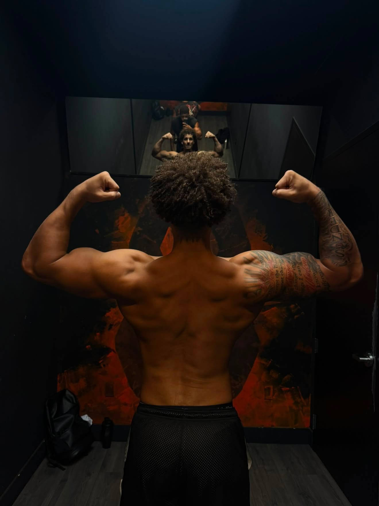 Eli Hughes flexing his back and arms in a gym mirror, tattoos visible, moody lighting