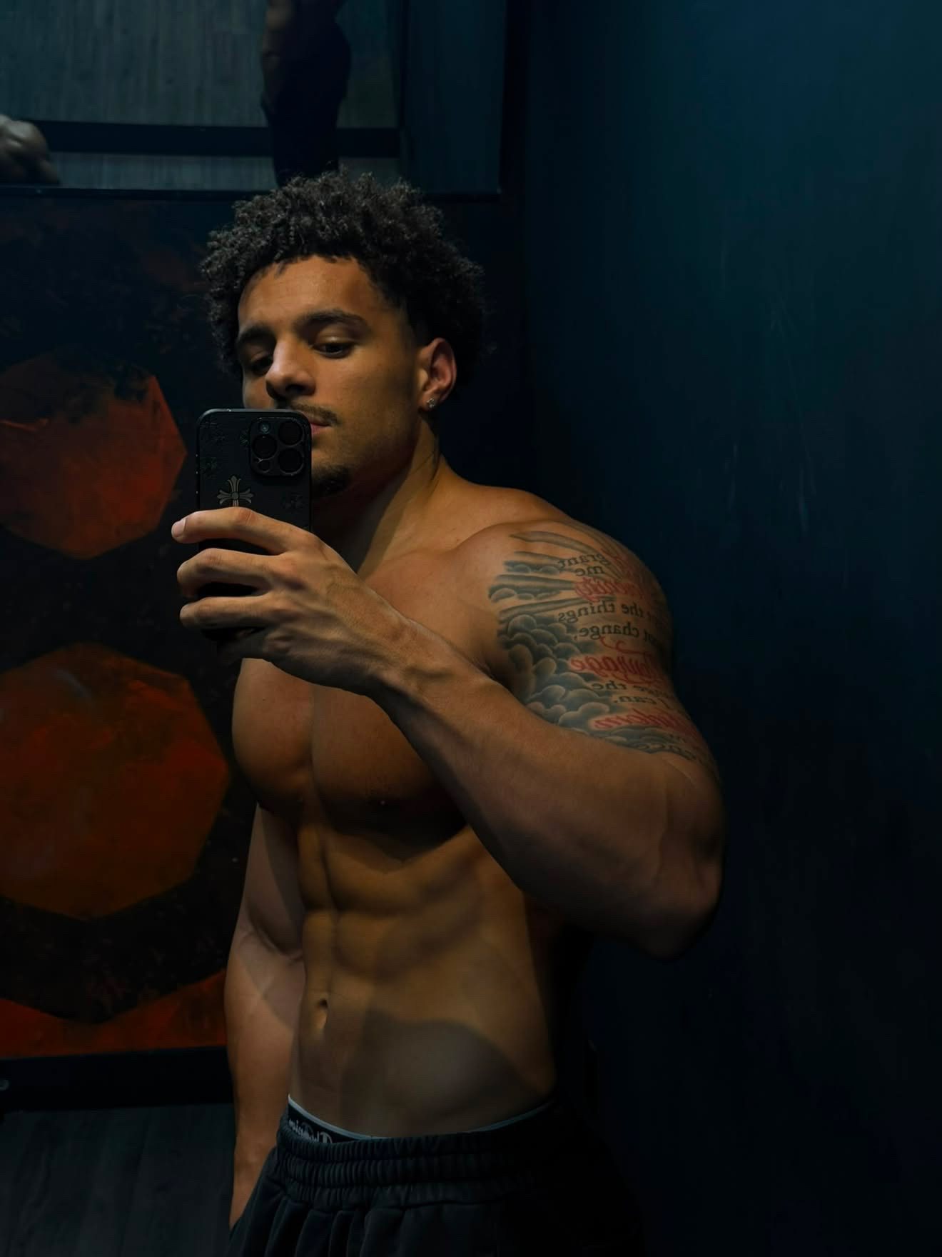 Eli Hughes taking a shirtless gym mirror selfie, showing his tattooed shoulder and physique