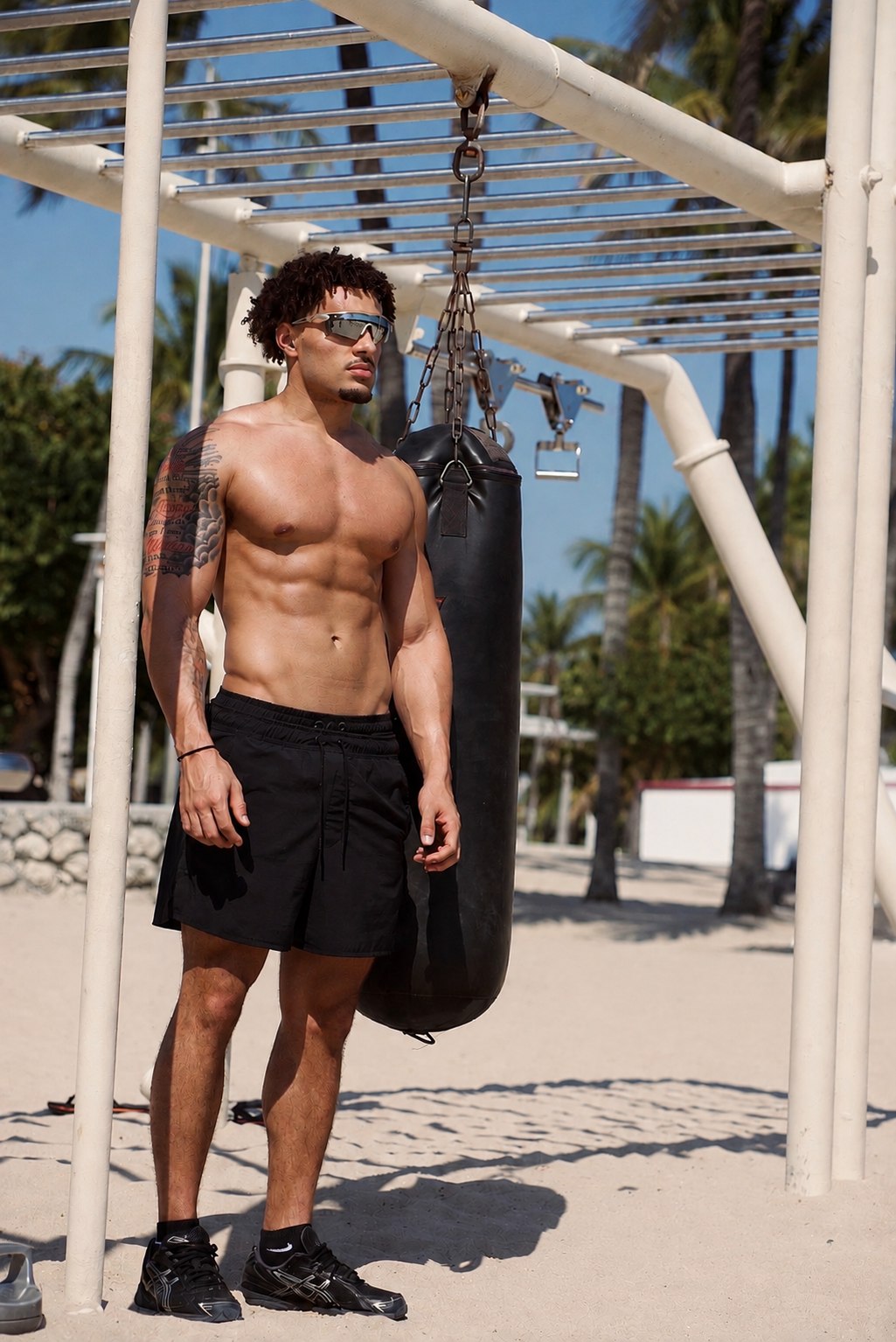 Eli Hughes standing shirtless on a Miami beach next to an outdoor punching bag rig, sunglasses on, palm trees behind him
